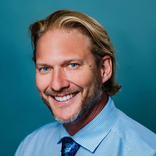 Dr. McNeill- Middle-aged man with long blonde hair smiling wearing a blue button down and blue tie